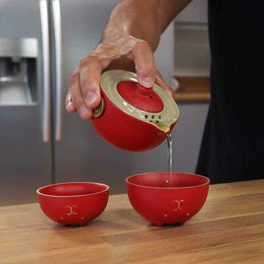 Red teapot pouring tea into two red cups on a wooden surface.
