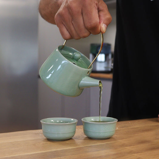 Person pouring tea from a green teapot into two cups on a wooden surface.